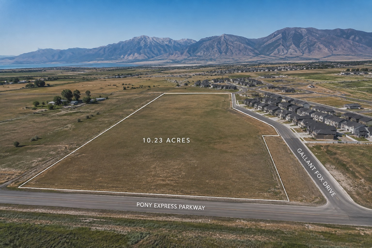 Aerial view of a 10.23-acre parcel on Pony Express Parkway, its boundary drawn in white, with a finished subdivision built to its edge and the Wasatch Range beyond.